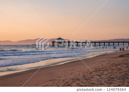 Manhattan Beach Pier extends into the Pacific Ocean at sunset in Los Angeles. A small building is silhouetted against the sky, with mountains in the distance. Manhattan Beach Pier extends into the Pacific Ocean at sunset in Los Angeles. A small building is silhouetted against the sky, with mountains in the distance. 124079364