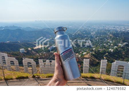 A person holds a metal water bottle in front of the Hollywood Sign, with Los Angeles cityscape visible in the distance, blending urban and natural elements. 124079382
