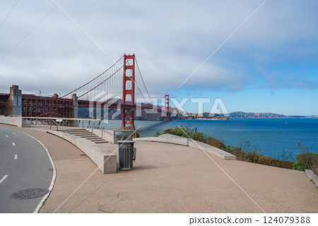 The Golden Gate Bridge spans San Francisco Bay, its International Orange color contrasting with the blue waters and partly cloudy sky. A pathway curves along the shoreline. 124079388