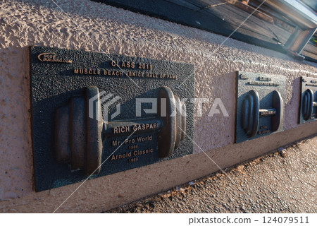 Plaque dedicated to Rich Gaspari at Muscle Beach Venice, Los Angeles, featuring a raised dumbbell design and casting shadows in sunlight. 124079511