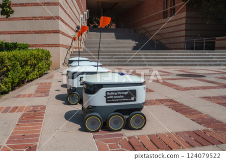 A group of delivery robots with orange flags line a brick walkway at UCLA. Steps and hedges frame the scene, showcasing technology in a university setting. 124079522