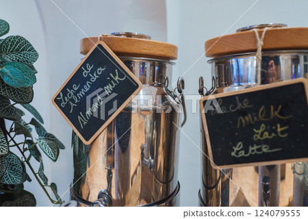 Two metal dispensers with wooden lids and chalkboard labels for almond milk and milk, set in a luxurious environment with a lush green plant. Two metal dispensers with wooden lids and chalkboard labels for almond milk and milk, set in a luxurious environment with a lush green plant. 124079555