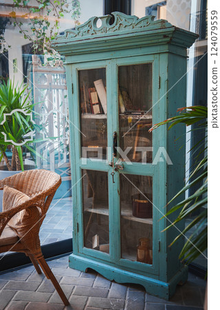 A vintage turquoise cabinet with glass doors displays books and decor, surrounded by lush plants and a wicker chair, in a Moroccan inspired setting. 124079559