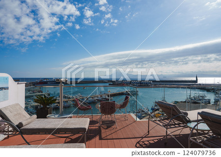 A terrace with lounge chairs and a telescope overlooks a marina filled with boats. A lighthouse is visible under a partly cloudy sky with bright sunlight. 124079736