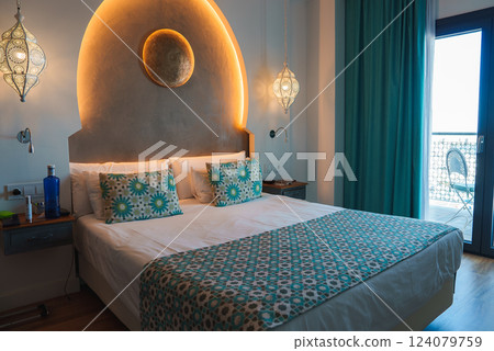 A hotel room features a large bed with patterned pillows, ornate lamps, and an arched headboard. A glass door opens to a balcony, enhancing the room's ambiance. A hotel room features a large bed with patterned pillows, ornate lamps, and an arched headboard. A glass door opens to a balcony, enhancing the room's ambiance. 124079759