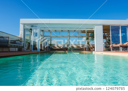 A rooftop pool area features modern Marrakech design, with sleek structures, large glass windows, seating, and lush plants under a clear blue sky. A rooftop pool area features modern Marrakech design, with sleek structures, large glass windows, seating, and lush plants under a clear blue sky. 124079785