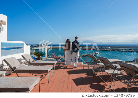 A couple stands on a terrace with sun loungers, overlooking a marina with docked boats. The scene includes a clear blue sky and distant mountains. 124079789