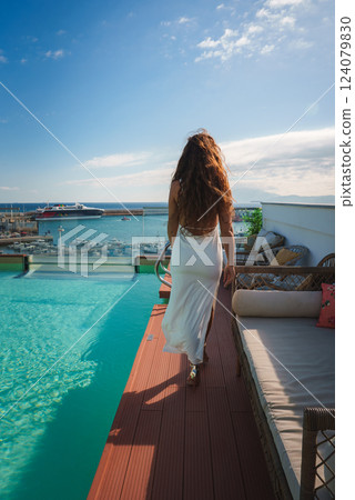 A woman in a white dress walks beside an infinity pool, with a marina and boats in the background. Clear skies and plush seating enhance the luxurious setting. A woman in a white dress walks beside an infinity pool, with a marina and boats in the background. Clear skies and plush seating enhance the luxurious setting. 124079830