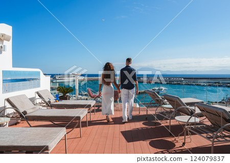 A couple walks hand in hand on a terrace with lounge chairs, overlooking a marina with yachts. Clear blue sky and distant mountains suggest a coastal setting. A couple walks hand in hand on a terrace with lounge chairs, overlooking a marina with yachts. Clear blue sky and distant mountains suggest a coastal setting. 124079837