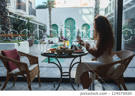 A woman enjoys a meal in a serene courtyard with a small pool, lush greenery, and arched doorways featuring intricate green tile work in Marrakech style. 124079880