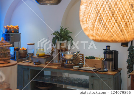 A luxury hotel interior features a wooden buffet with breakfast items, including a juice dispenser, juicer, and oranges. A woven pendant light adds warmth. A luxury hotel interior features a wooden buffet with breakfast items, including a juice dispenser, juicer, and oranges. A woven pendant light adds warmth. 124079896