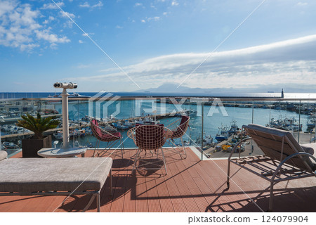 A luxurious terrace with modern furniture and a telescope overlooks a bustling marina filled with boats and yachts, set against a mountainous landscape. 124079904
