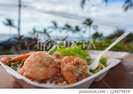 A plate of garlic shrimp over rice with greens on a wooden table, set against palm trees, ocean views, parked cars, and a blue sky in Hawaii. A plate of garlic shrimp over rice with greens on a wooden table, set against palm trees, ocean views, parked cars, and a blue sky in Hawaii. 124079934