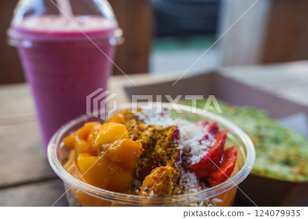 A vibrant acai bowl with mango, strawberries, coconut, and granola sits next to a pink smoothie. Blurred greenery suggests an outdoor Hawaii cafe setting. A vibrant acai bowl with mango, strawberries, coconut, and granola sits next to a pink smoothie. Blurred greenery suggests an outdoor Hawaii cafe setting. 124079935
