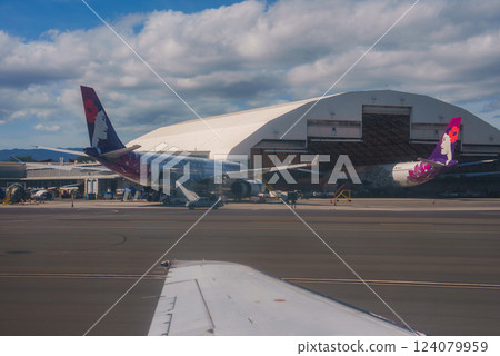 View of two Hawaiian Airlines planes parked near a white hangar, with mountains in the background and a partly cloudy sky, seen from another aircraft. 124079959
