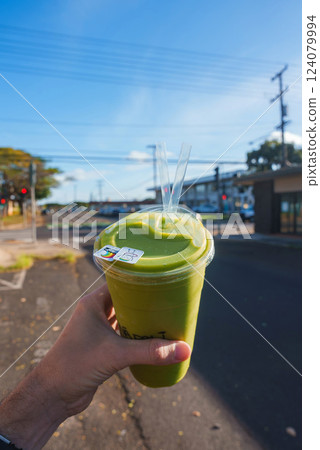 A hand holds a green smoothie in a clear cup with two straws, set against a sunny street with traffic lights, power lines, and small buildings. 124079994