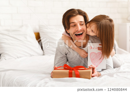 Little girl celebrating Father's Day with daddy, daughter presenting gift box and card, kissing dad from behind, bedroom interior Little girl celebrating Father's Day with daddy, daughter presenting gift box and card, kissing dad from behind, bedroom interior 124080844