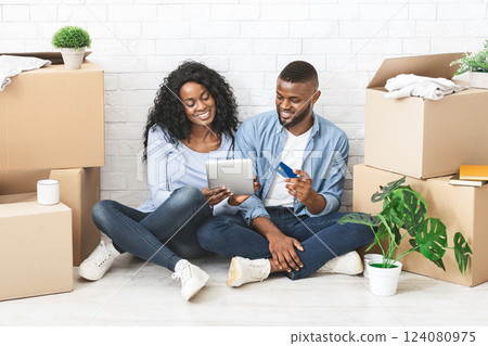 Young black couple buying furniture online, using digital tablet and credit card, sitting on floor in new apartment 124080975
