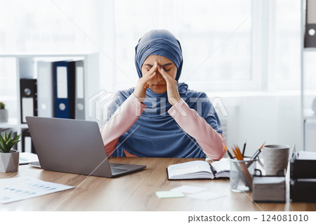 Fatigue Concept. Exhausted muslim busineswoman sitting at desk and touching her nose bridge, suffering from migraine and overworking in office 124081010