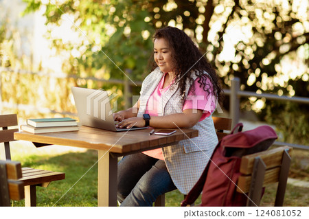 A student is engaged with a laptop outdoors, surrounded by greenery, enjoying the sun while studying. Books are stacked beside her, and a backpack rests on the chair. A student is engaged with a laptop outdoors, surrounded by greenery, enjoying the sun while studying. Books are stacked beside her, and a backpack rests on the chair. 124081052