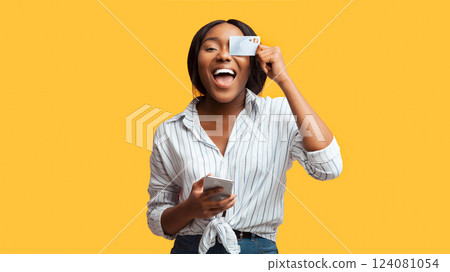 Cheerful Afro Girl Holding Smartphone And Credit Card Covering Eye Having Fun Posing Over Pink Studio Background. Panorama Cheerful Afro Girl Holding Smartphone And Credit Card Covering Eye Having Fun Posing Over Pink Studio Background. Panorama 124081054