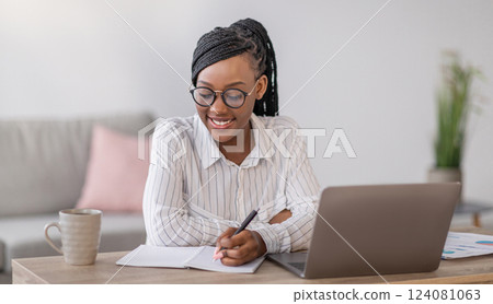 Positive black business woman with stylish glasses manager working from home during COVID-19 pandemic, using modern laptop and taking notes, sitting at workdesk in living room, copy space 124081063