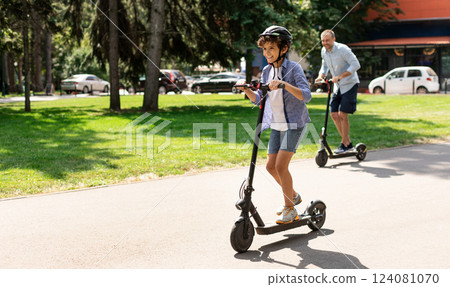 Happy Loving Family. Full body length photo of excited curly boy in protective helmet driving motorized push scooter with daddy at park, spending time together outdoors at summer, selective focus 124081070
