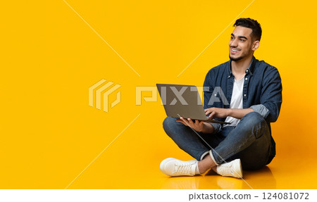 Happy arab young man sitting on floor, using new modern laptop, typing on keyboard and smiling over yellow studio background, looking at copy space for text or advertisement, panorama 124081072