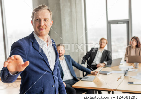 A businessman in a blue suit stands and addresses his colleagues who are sitting at a conference table during a business presentation. A businessman in a blue suit stands and addresses his colleagues who are sitting at a conference table during a business presentation. 124081295