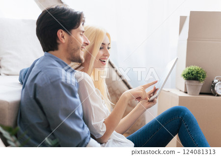 Cheerful Couple Using Tablet Computer Sitting Among Moving Boxes After Relocation To New Place. Empty Space 124081313