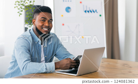 Young ceo portrait. Confident millennial african american businessman sitting in office with smartphone and laptop, smiling and posing to camera Young ceo portrait. Confident millennial african american businessman sitting in office with smartphone and laptop, smiling and posing to camera 124081362