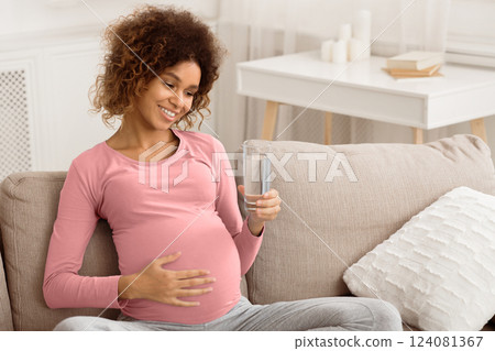 Pregnancy and hydration. Happy woman holding glass of water, sitting on couch at home, free space Pregnancy and hydration. Happy woman holding glass of water, sitting on couch at home, free space 124081367