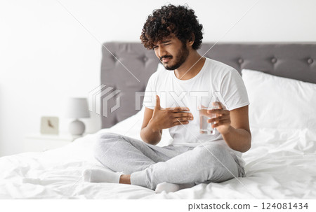 Unhappy millennial indian guy suffering from heartburn, waking up in morning, sitting on bed and holding glass of water, wearing pajamas, copy space. Acid reflux, pain in stomach concept 124081434