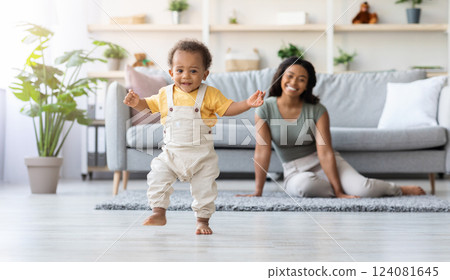 First Steps. Adorable Black Infant Child Walking In Living Room At Home, Cute African American Toddler Boy Stepping On Floor And Looking At Camera, His Proud Mother Smiling On Background, Free Space First Steps. Adorable Black Infant Child Walking In Living Room At Home, Cute African American Toddler Boy Stepping On Floor And Looking At Camera, His Proud Mother Smiling On Background, Free Space 124081645