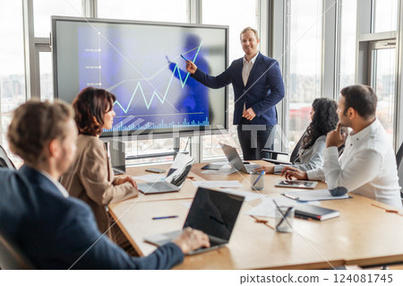 A business professional stands near a large digital screen showing an upward trending line graph. The presenter gestures towards the screen as they explain the chart to a group of colleagues A business professional stands near a large digital screen showing an upward trending line graph. The presenter gestures towards the screen as they explain the chart to a group of colleagues 124081745