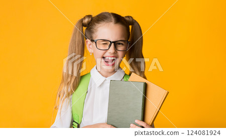 Funny Elementary School Girl Winking At Camera Holding Books Posing Over Yellow Background In Studio. Happy Back-To-School Funny Elementary School Girl Winking At Camera Holding Books Posing Over Yellow Background In Studio. Happy Back-To-School 124081924