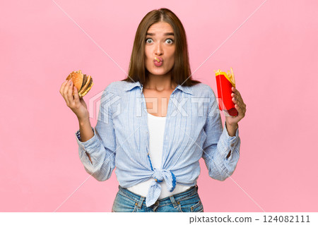 Oops. Surprised Woman Eating Unhealthy Burger And French Fries Standing On Yellow Background. Studio Shot Oops. Surprised Woman Eating Unhealthy Burger And French Fries Standing On Yellow Background. Studio Shot 124082111