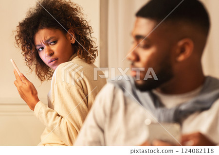Afro woman suspecting husband in betrayal, using phone and looking at man 124082118