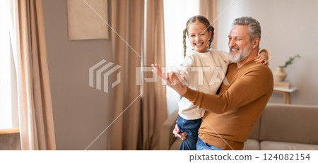 A joyful grandfather lifts his smiling granddaughter in a cozy indoor space filled with natural light. The warmth of their bond is evident as they share a playful moment together. A joyful grandfather lifts his smiling granddaughter in a cozy indoor space filled with natural light. The warmth of their bond is evident as they share a playful moment together. 124082154