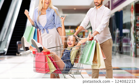 A young girl sits in a shopping cart, being pushed by her parents. The girl is smiling and laughing as she holds onto the handle of the cart. Her parents are also smiling and look happy 124082215
