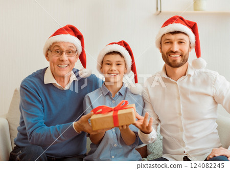 Christmas Male Family Portrait. Grandfather, Adult Son And Grandson In Santa Hats Holding Xmas Gift Sitting On Sofa At Home. 124082245