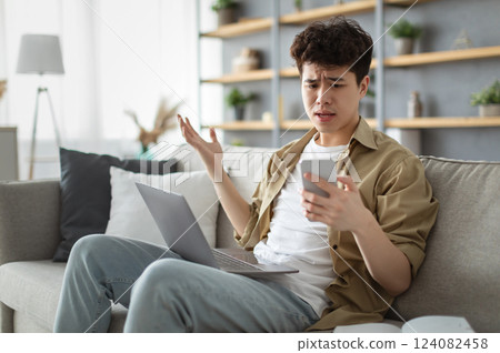Confused asian man looking at phone screen, sitting on couch with computer on lap in living room, upset frustrated male reading bad news on message, having problem with device or Internet connection Confused asian man looking at phone screen, sitting on couch with computer on lap in living room, upset frustrated male reading bad news on message, having problem with device or Internet connection 124082458