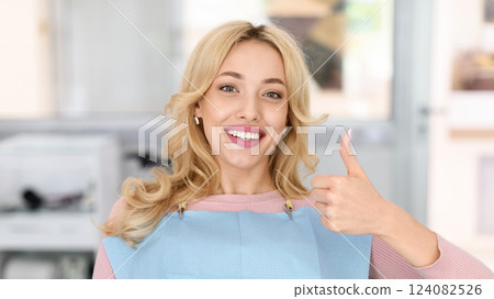 Pretty blonde lady showing her beautiful healthy smile and thumb up, panorama with copy space. Female patient of modern dental clinic sitting in chair, recommending her dentist, closeup 124082526