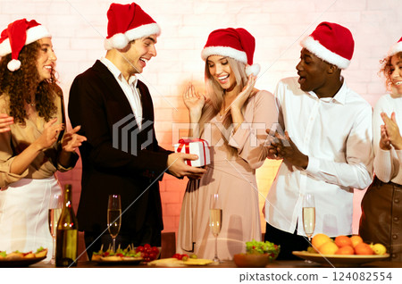 Christmas Party Concept. Friends Congratulating Girl Giving Her Xmas Gift Celebrating Winter Holidays At Home Christmas Party Concept. Friends Congratulating Girl Giving Her Xmas Gift Celebrating Winter Holidays At Home 124082554
