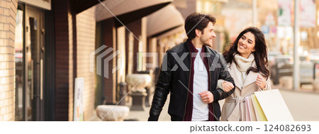 A couple walks together, smiling and holding shopping bags, dressed in casual attire, enjoying their time on a sunny day, copy space A couple walks together, smiling and holding shopping bags, dressed in casual attire, enjoying their time on a sunny day, copy space 124082693