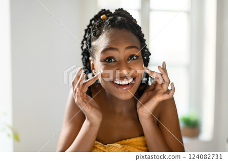 A young black woman with braided black hair smiles brightly while applying moisturizer to her face. She is standing in a bathroom with a white tiled wall and a window in the background. 124082731