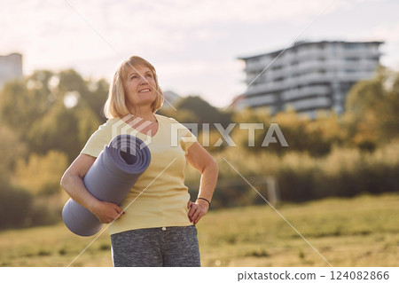 Standing and holding yoga mat. Senior woman having nice weekend outdoors on the field at sunny day Standing and holding yoga mat. Senior woman having nice weekend outdoors on the field at sunny day 124082866