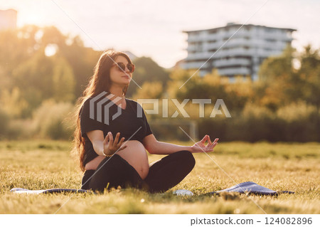 Young beautiful pregnant woman that is in sunglasses is doing yoga on the mat outdoors on the field 124082896