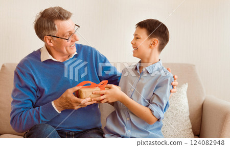 Grandpa's Birthday. Cheerful Boy Congratulating Grandfather Giving Him Wrapped Gift Box Sitting On Sofa Indoor. Selective Focus Grandpa's Birthday. Cheerful Boy Congratulating Grandfather Giving Him Wrapped Gift Box Sitting On Sofa Indoor. Selective Focus 124082948