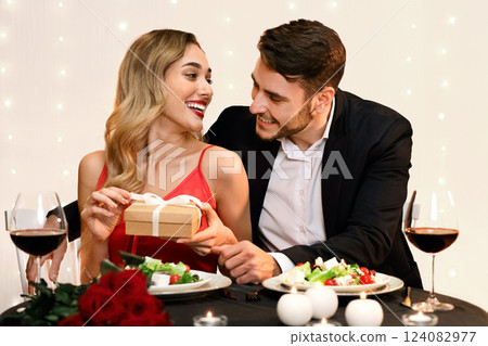 Loving man giving surprise gift for valentine's day to his girlfriend, happy woman unwrapping present on dinner in restaurant, free space Loving man giving surprise gift for valentine's day to his girlfriend, happy woman unwrapping present on dinner in restaurant, free space 124082977